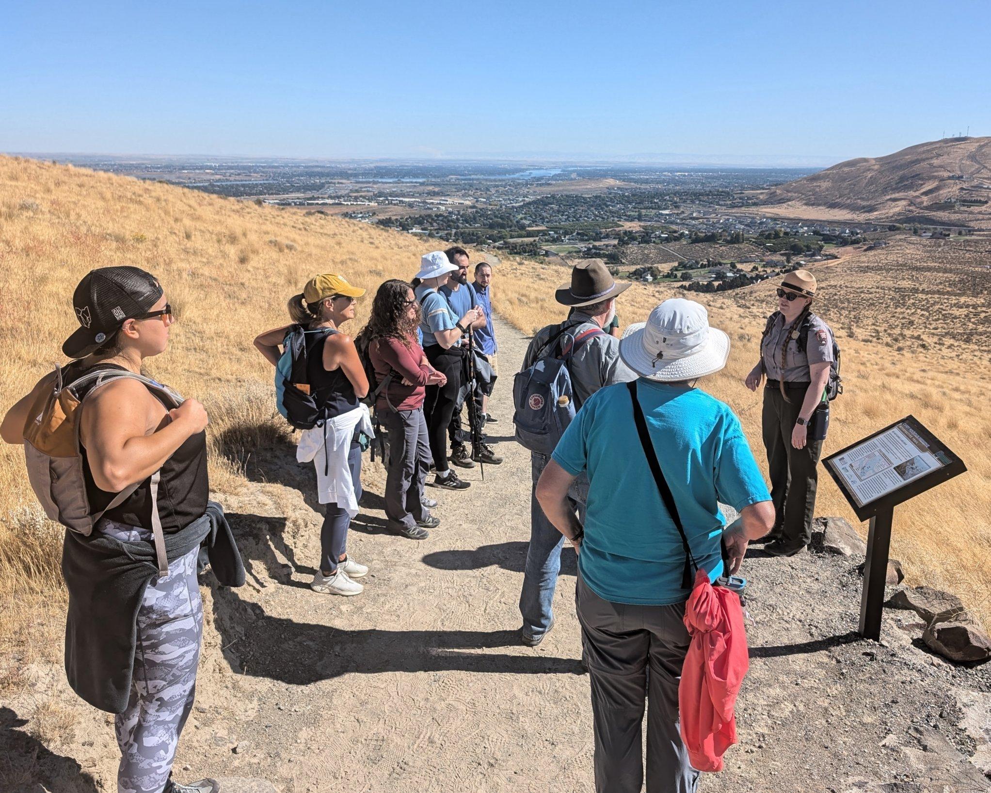 Uniformed ranger addresses 8 people on a gravel trail with dry brush on either side.