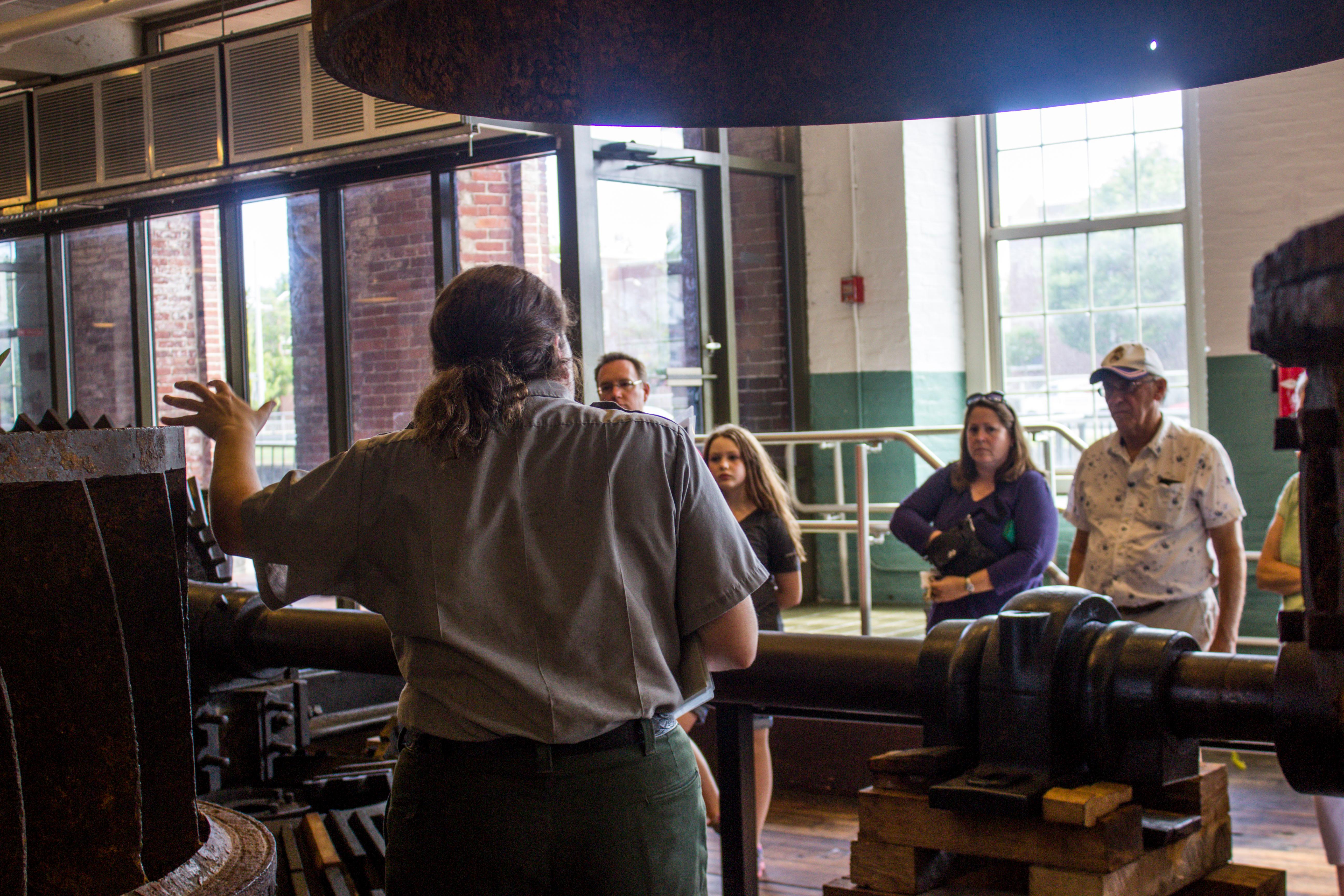 A ranger gestures towards a deconstructed historic turbine while speaking to a group of visitors