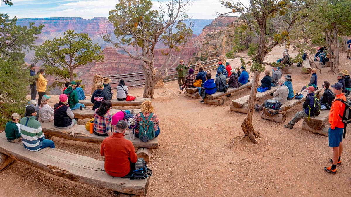 in an outdoor amphitheater, people are sitting on log benches and listening to a park ranger talk.