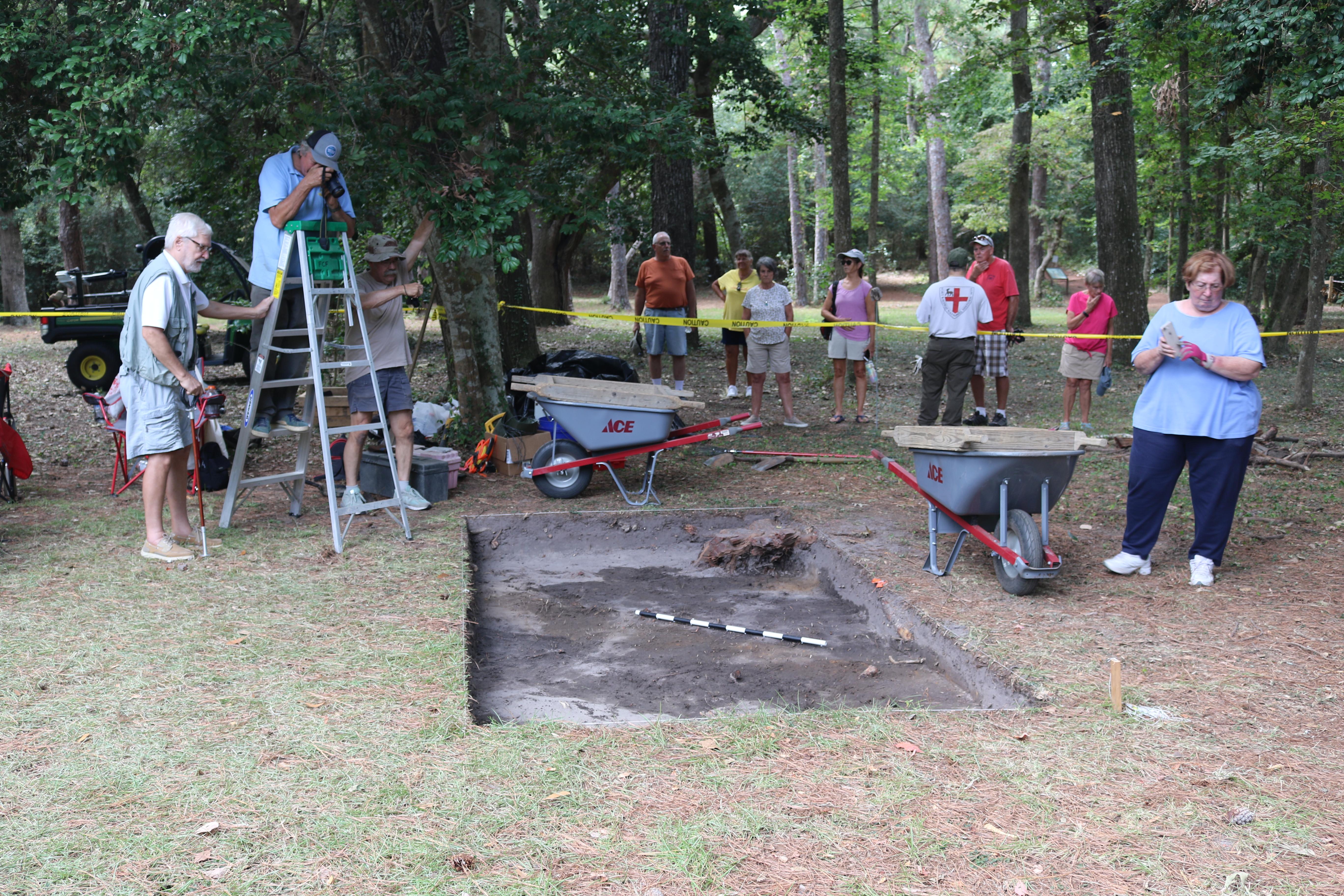 A group of people around a shallow pit with tools scattered around them.