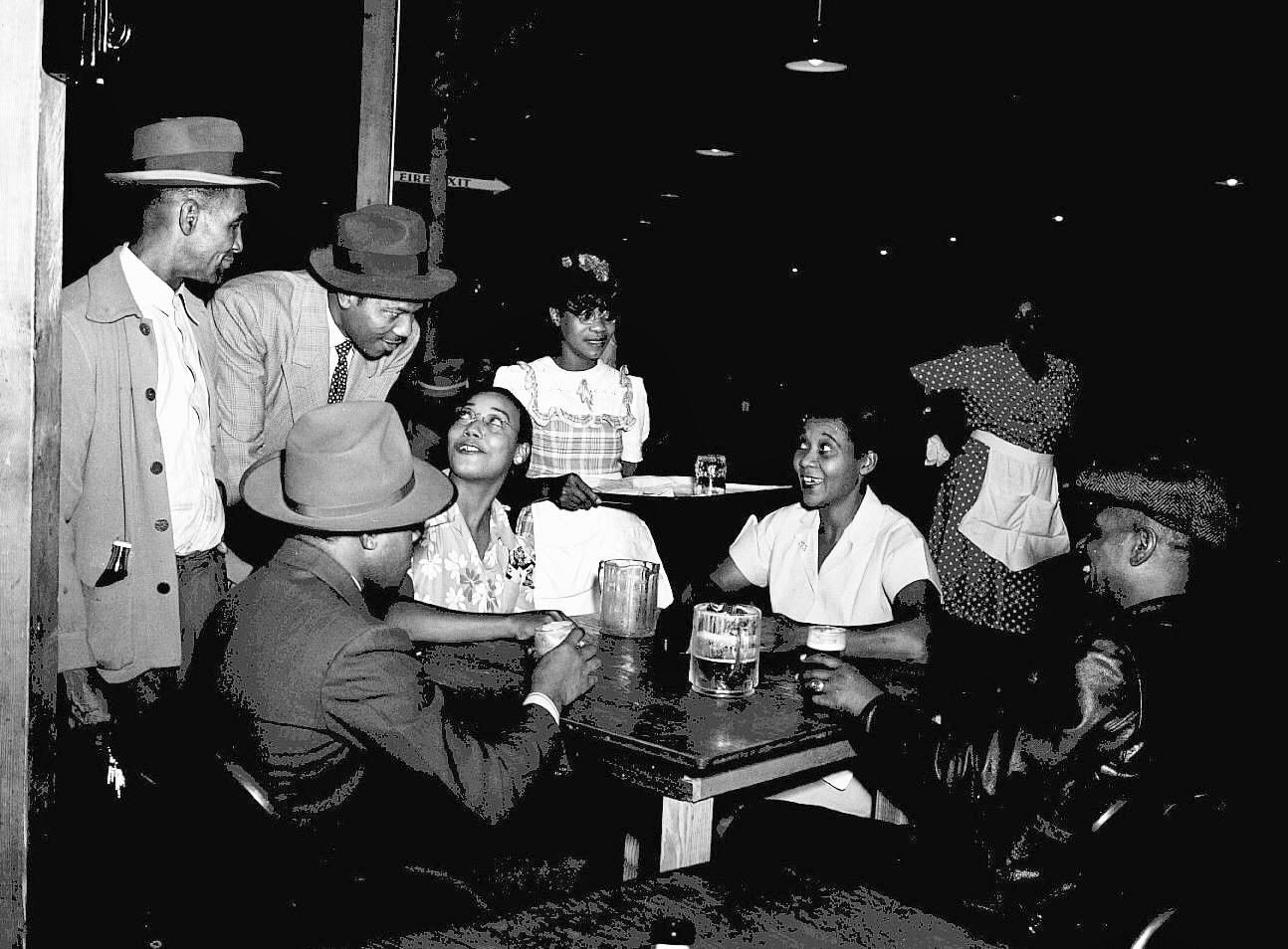 4 African Americans seated at a restaurant table talking with 4 people standing around the table.