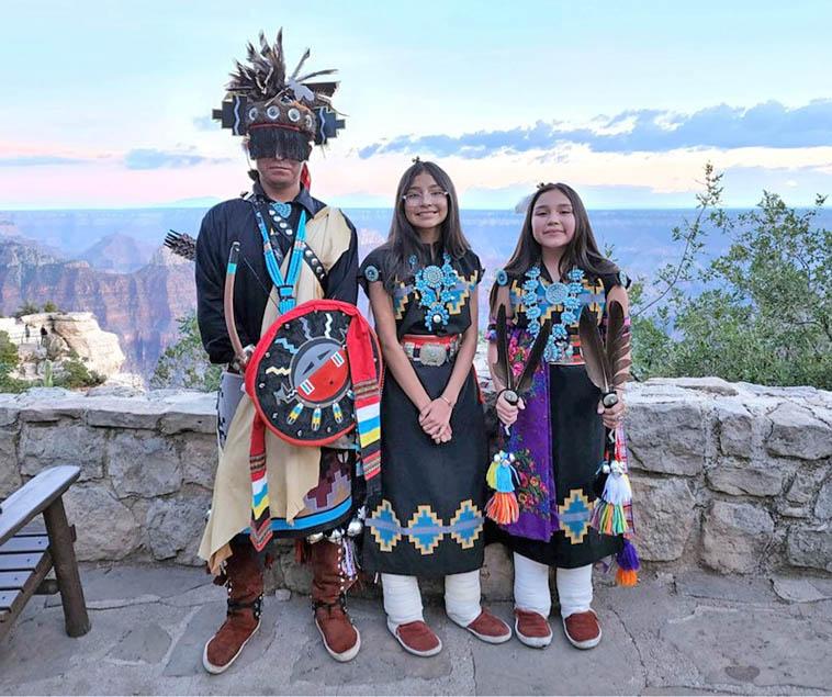 3 young Native American dancers, wearing ceremonial clothes, are posing for a group photo outdoors.