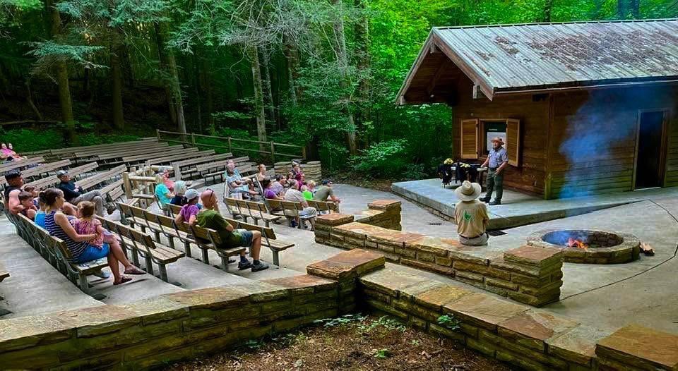 a group of people sitting on benches listening to a ranger speak on stage