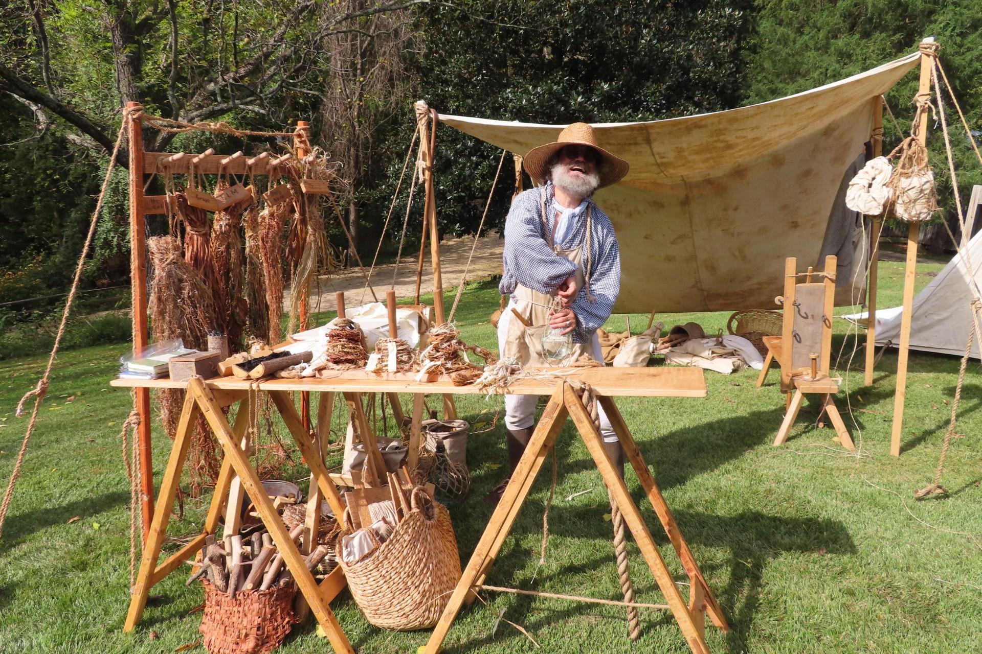 A man dressed in colonial-era clothes stands outside behind a wooden bench filled with historical cr