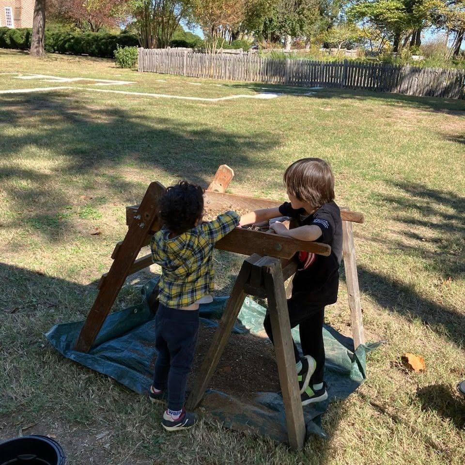 Two children standing in front of a standing wooden sand sifter. In the background is a sunny field.