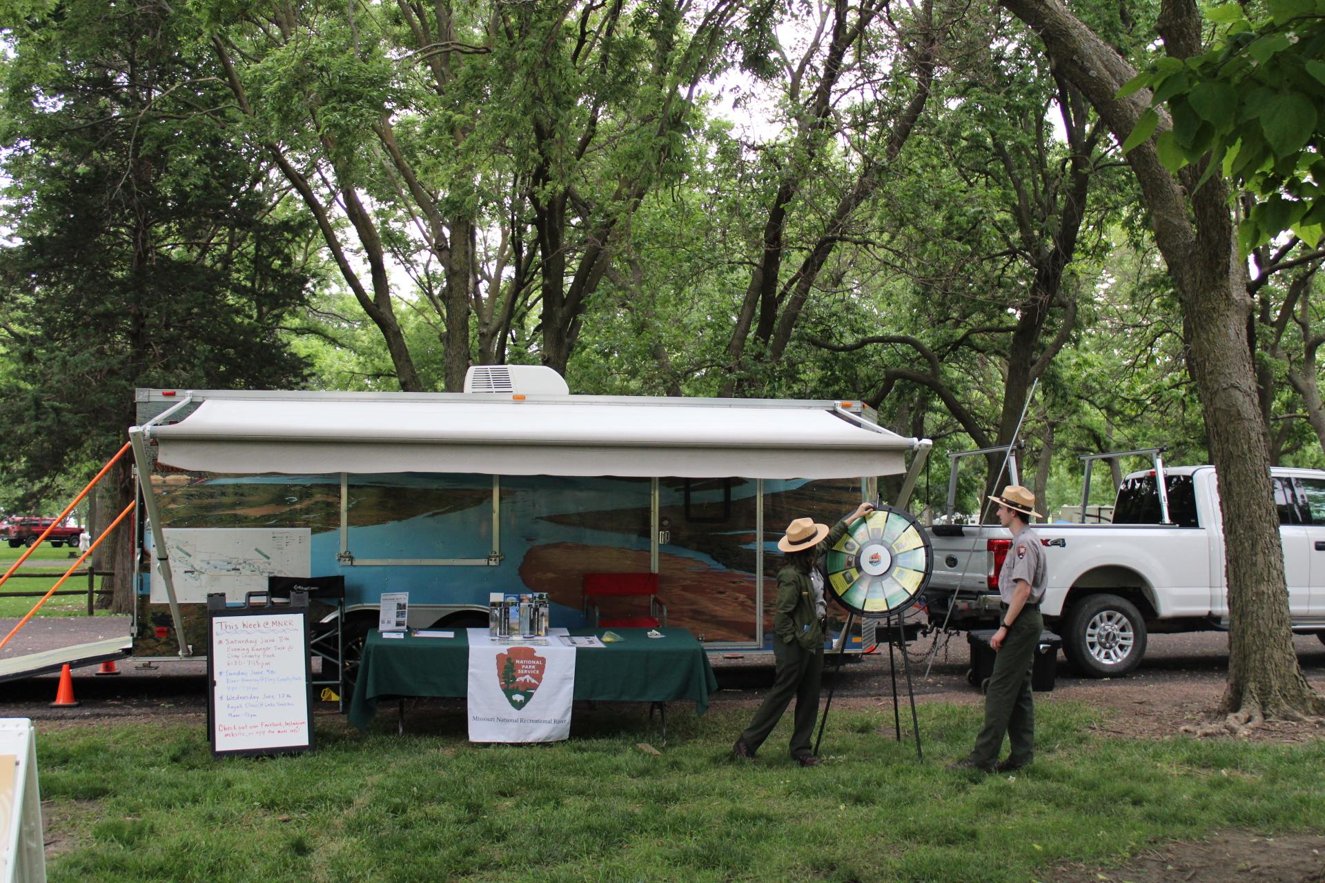 Two park rangers stand by a large trailer in an area with many trees.