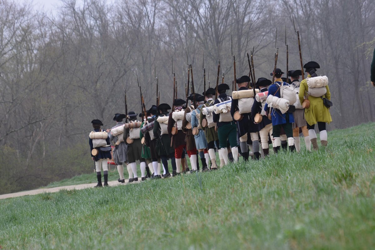 A column of Militia soldiers march down the dirt road surrounded by trees