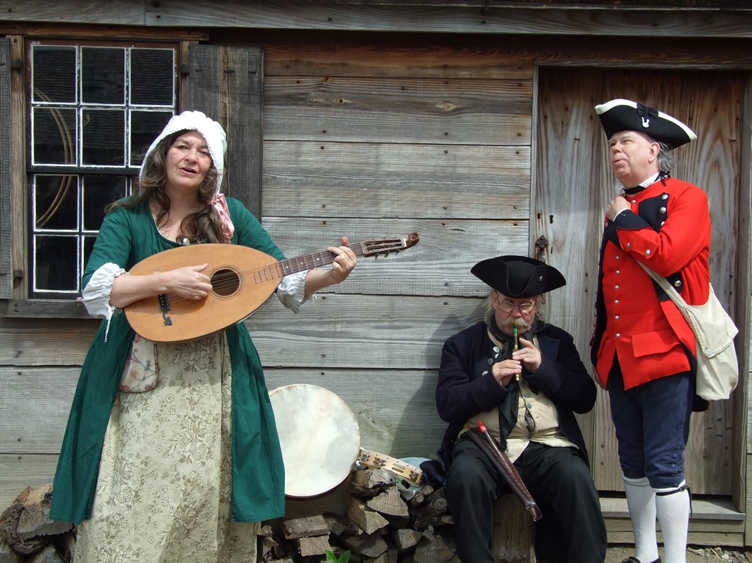 A woman stands playing a guitar-like instrument while a man stands and sings nearby.
