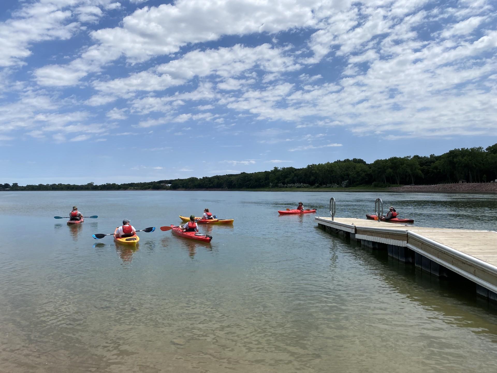 A group of people in kayaks float on a river near a boat dock on a clear day