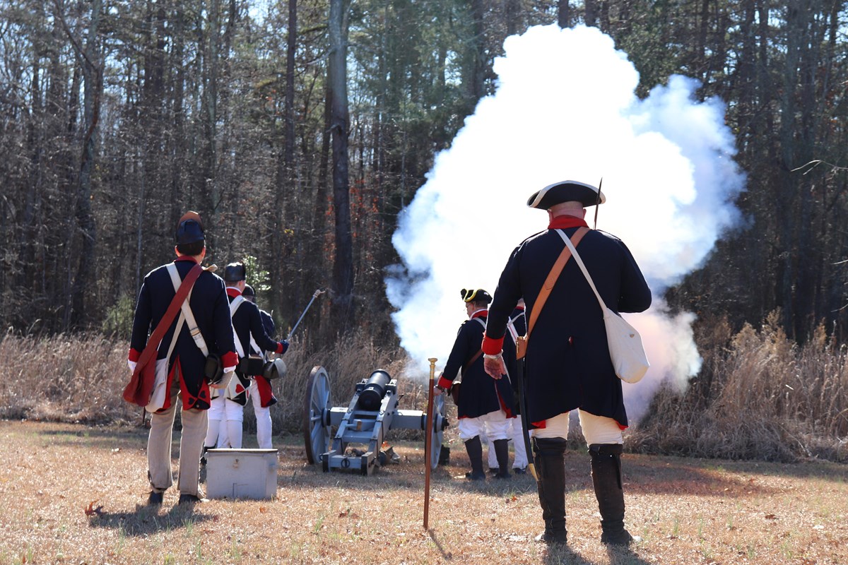 Reenactors in colonial uniform fire reproduction cannon. White smoke rises in foreground.
