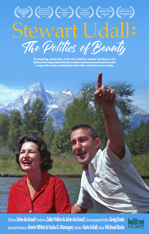 A woman and a man sit in front of a lake with blue skies, trees, and mountains in the background.