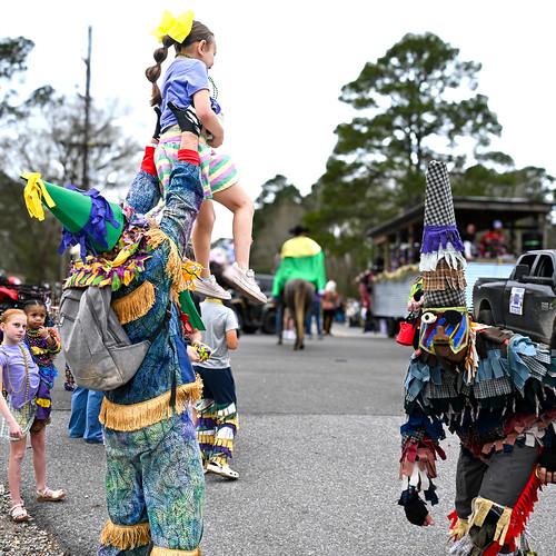To Country Mardi Gras participants play in the road in costume. One has lifted up a child.