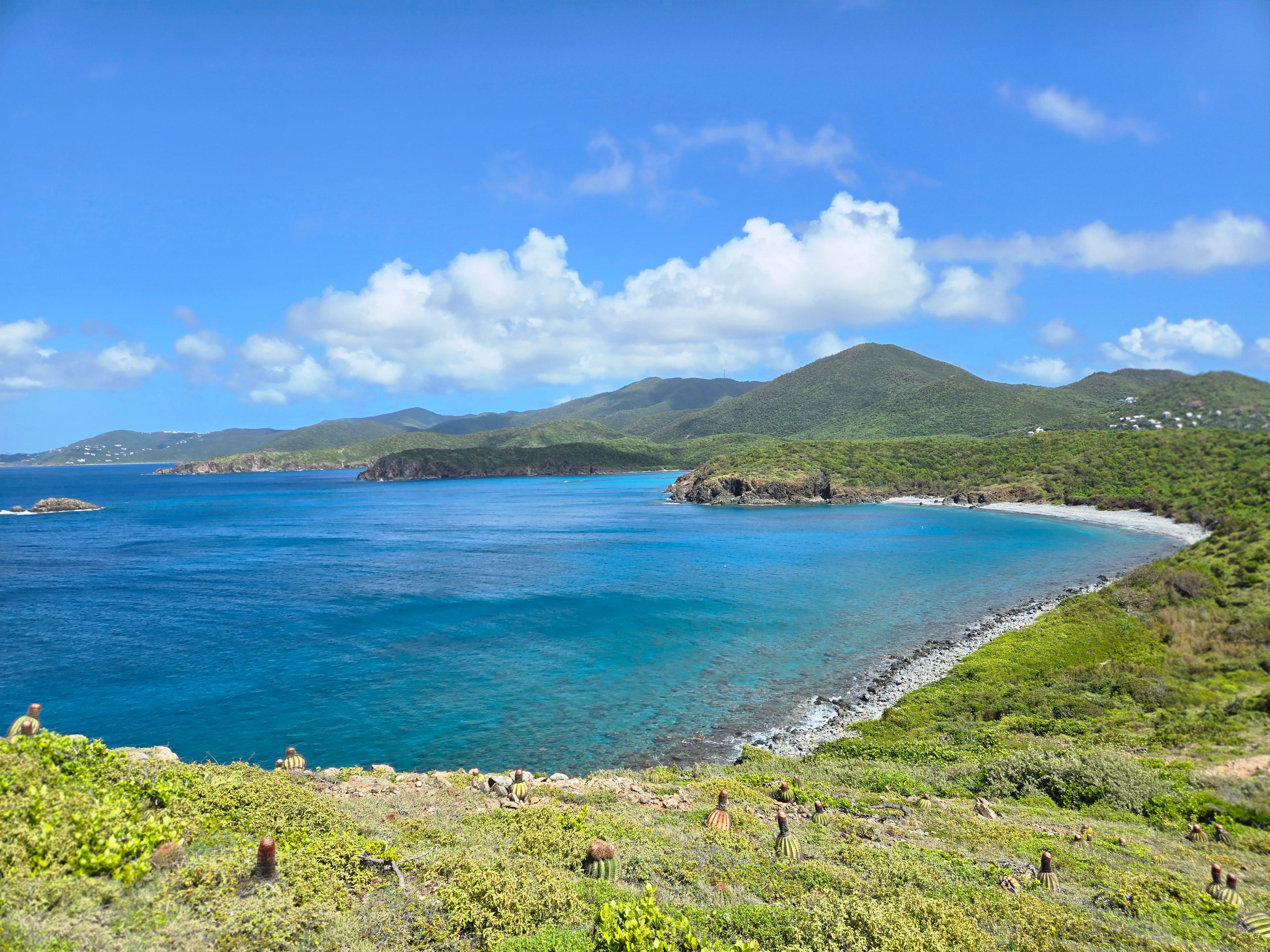 Image of a trail and beach with a cloudy blue sky, green hills, and blue water.
