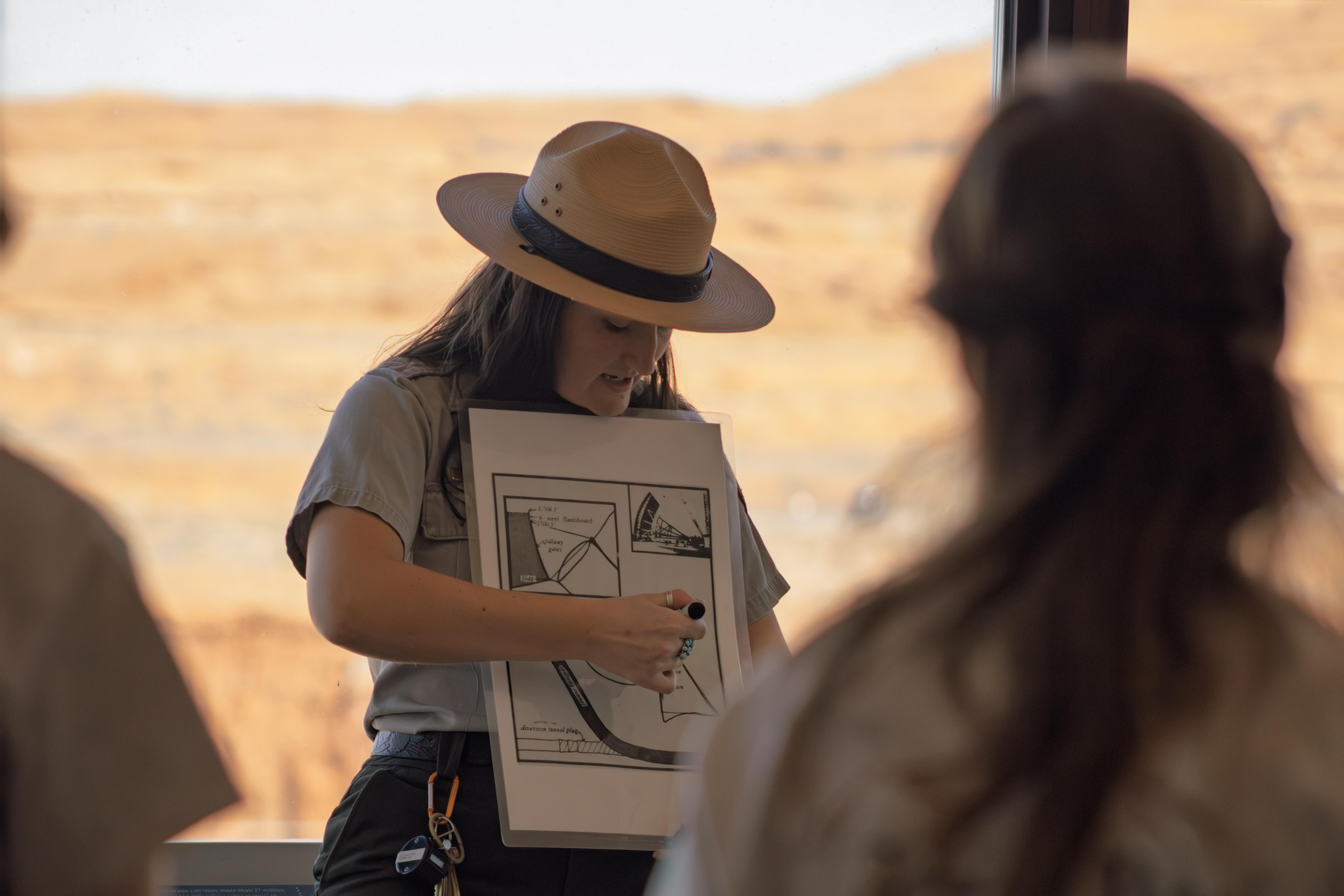 A park ranger points to a diagram of a dam in front of a group of people.