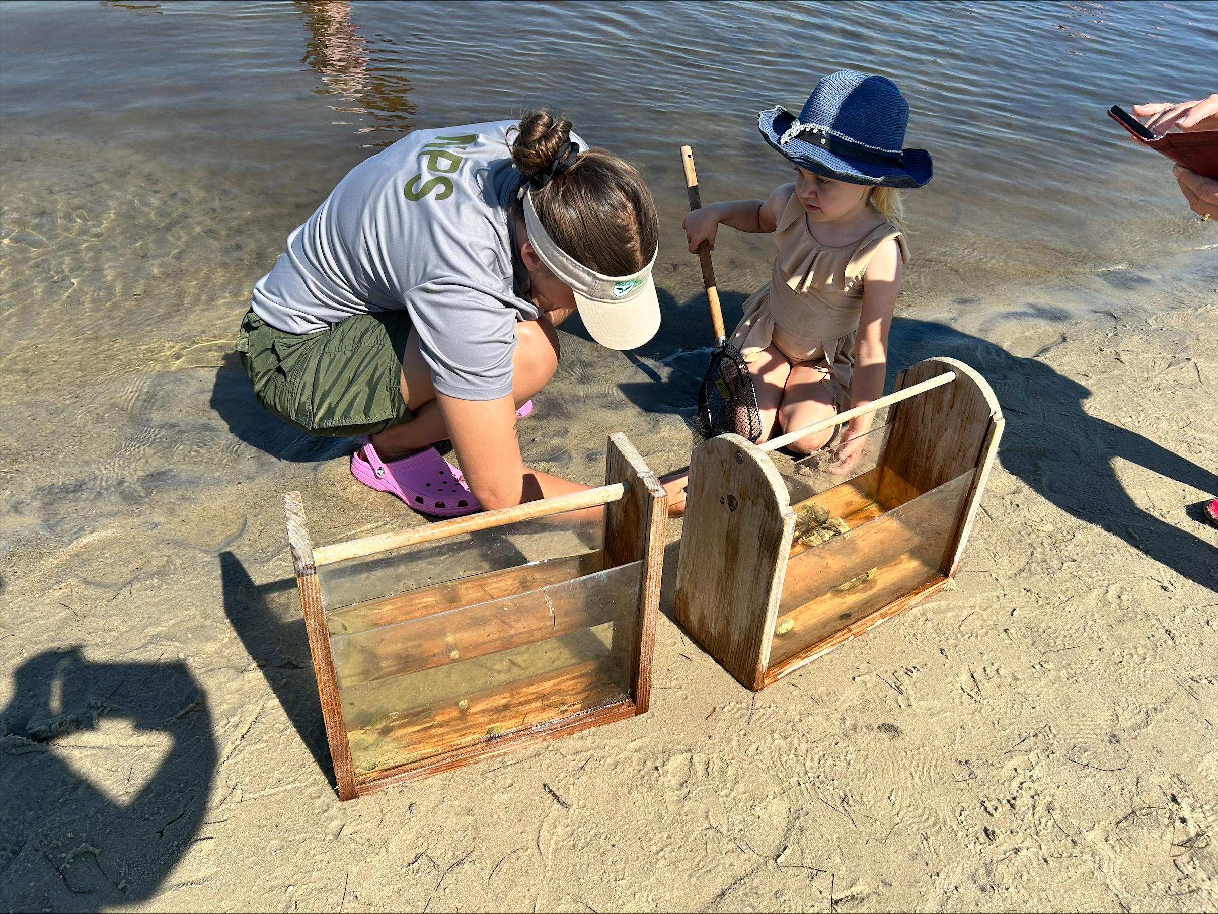 Ranger and child kneel on beach observing something.