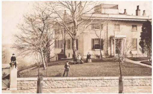 A sizable house sits on a raised plot in winter. Two boys can be seen on a stone fence.