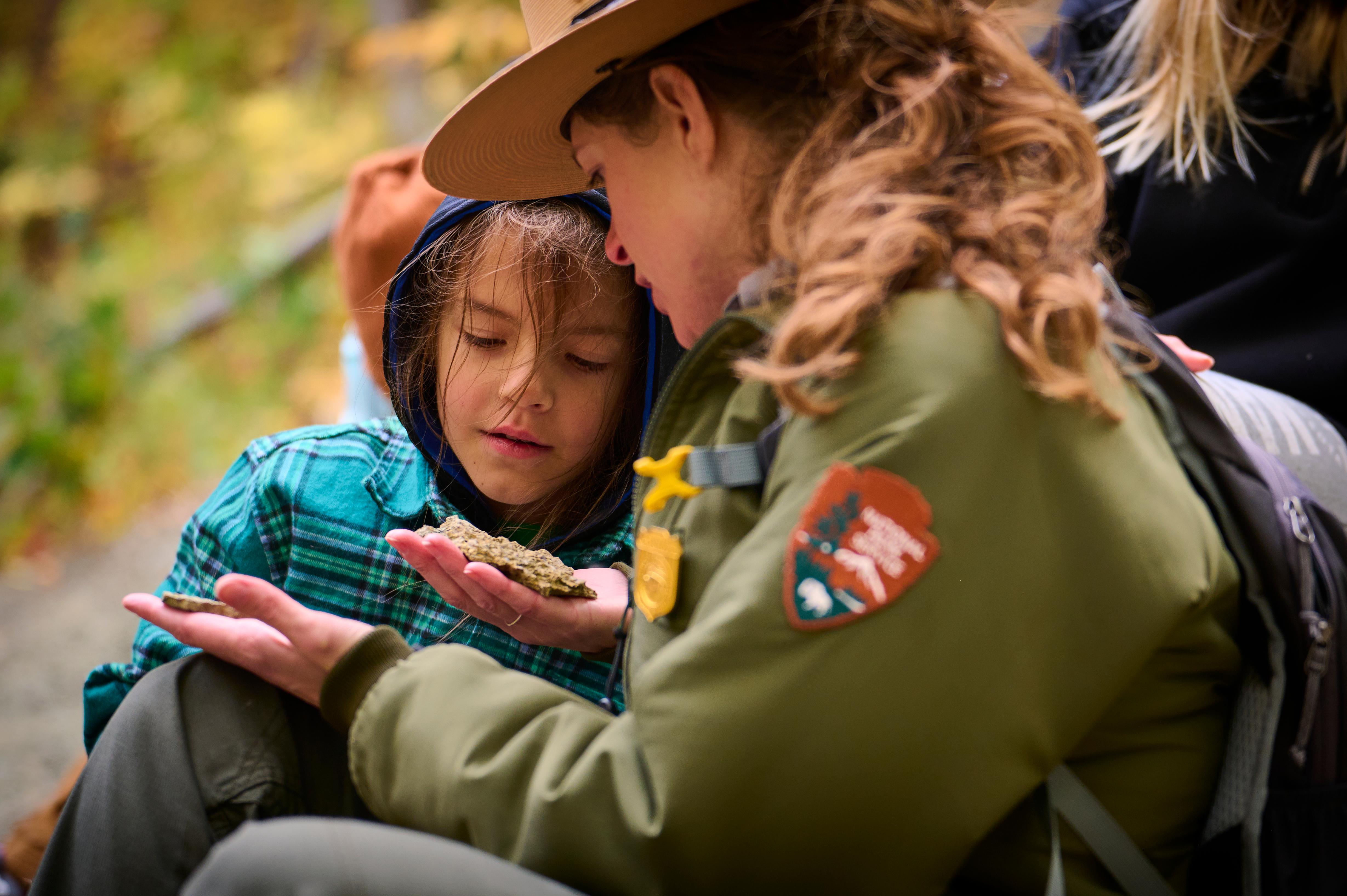 A park ranger sits with a young child to look at fossils in their hand.