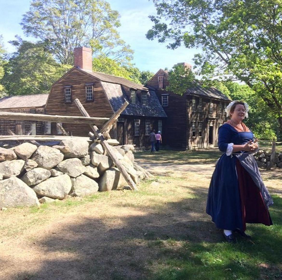 A Park Ranger in 18th Century women's dress stands in front of a large wood building and stone wall.