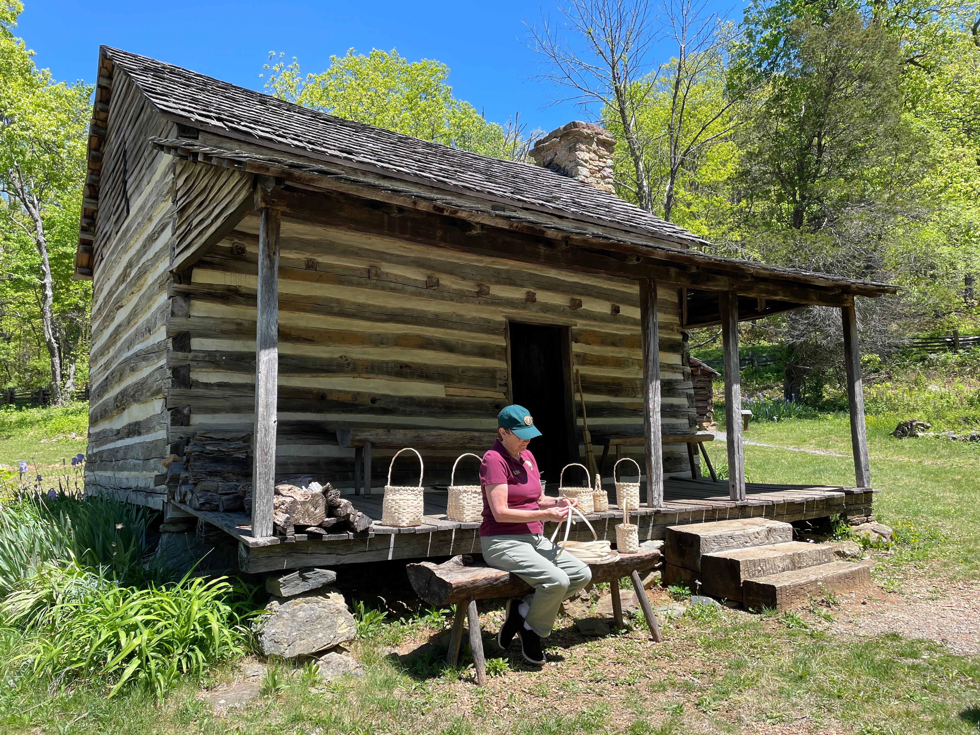 A woman sits on a bench outside a log cabin surrounded by 6 woven baskets.