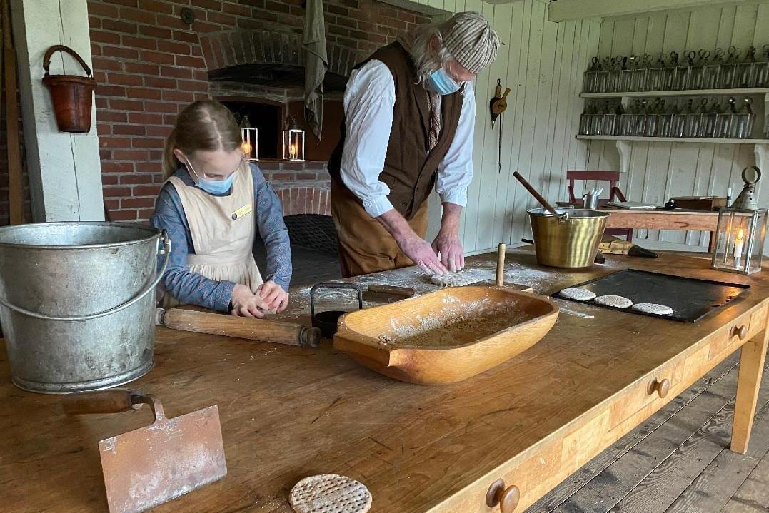 Two costumed interpreters - a young girl and an adult man - roll dough indoors.