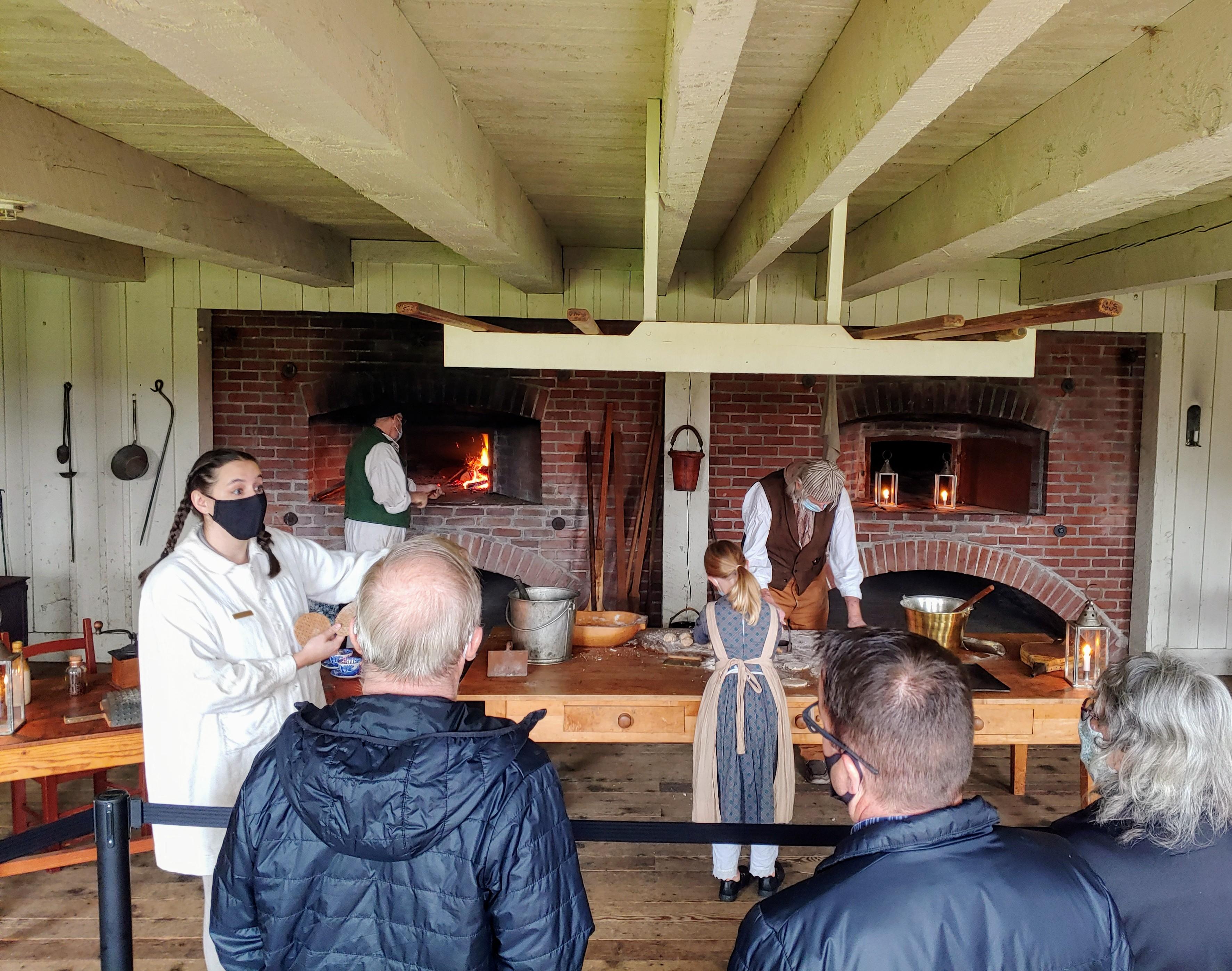 Visitors in foreground view baking demonstration in background. Volunteers roll and bake dough.