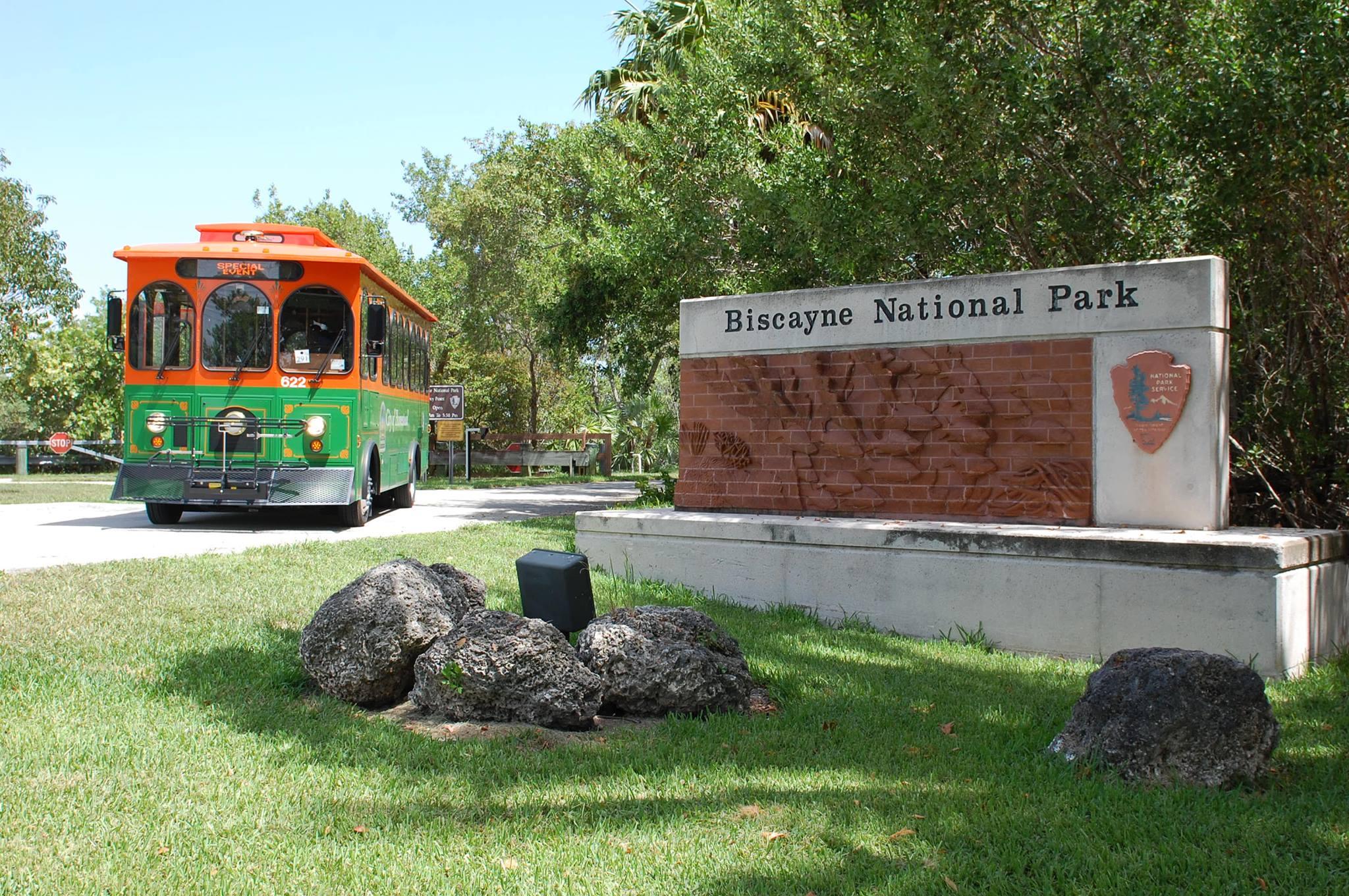 A green and orange trolley driving on a road next to a sign that says 'Biscayne National Park'