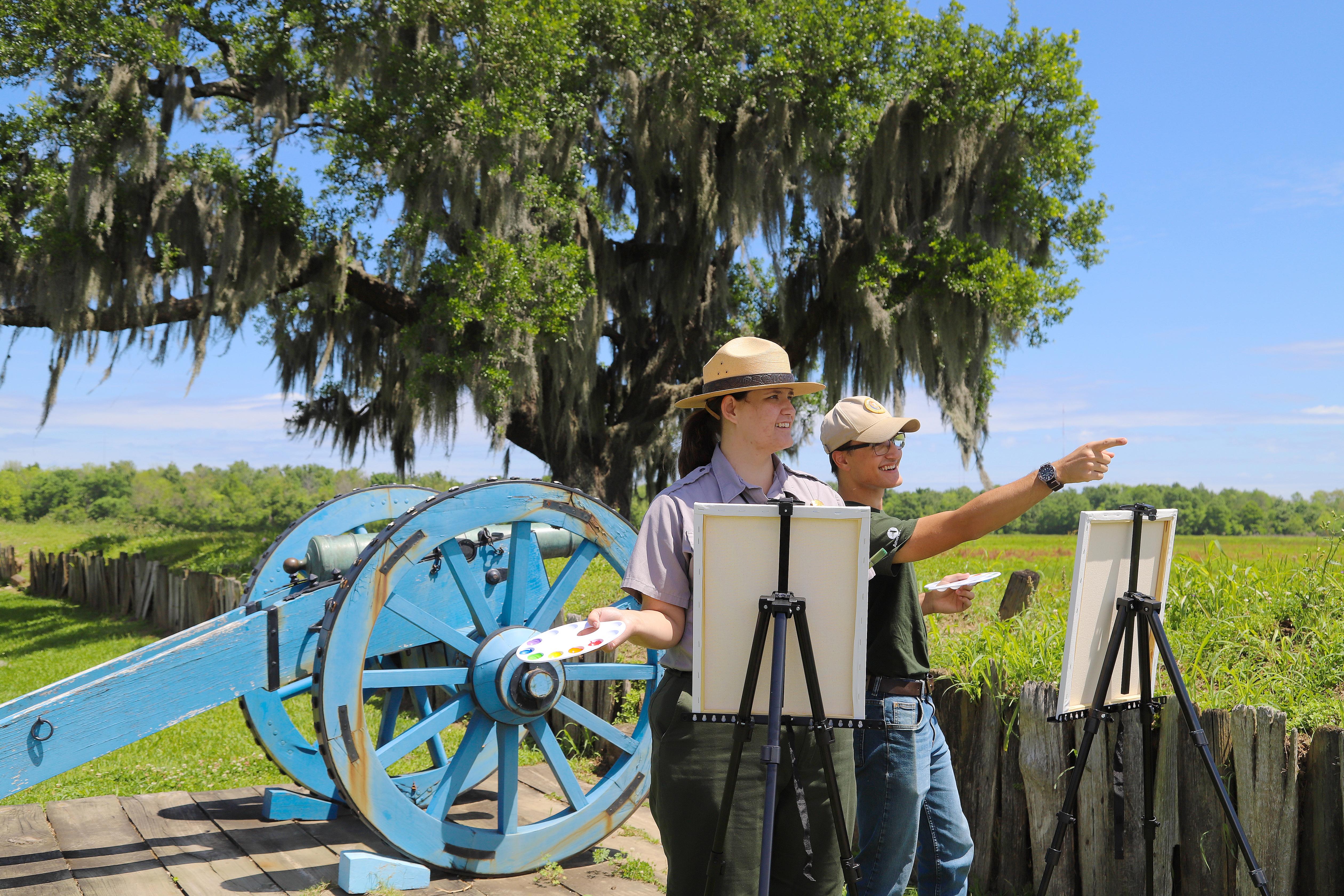 Two rangers stand behind canvases on easels outside on a sunny day. One ranger points at a distant s