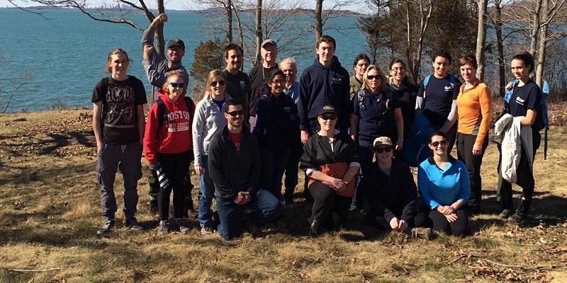 Three rows of people lined up for a photo in a grassy area with trees and the harbor in background.