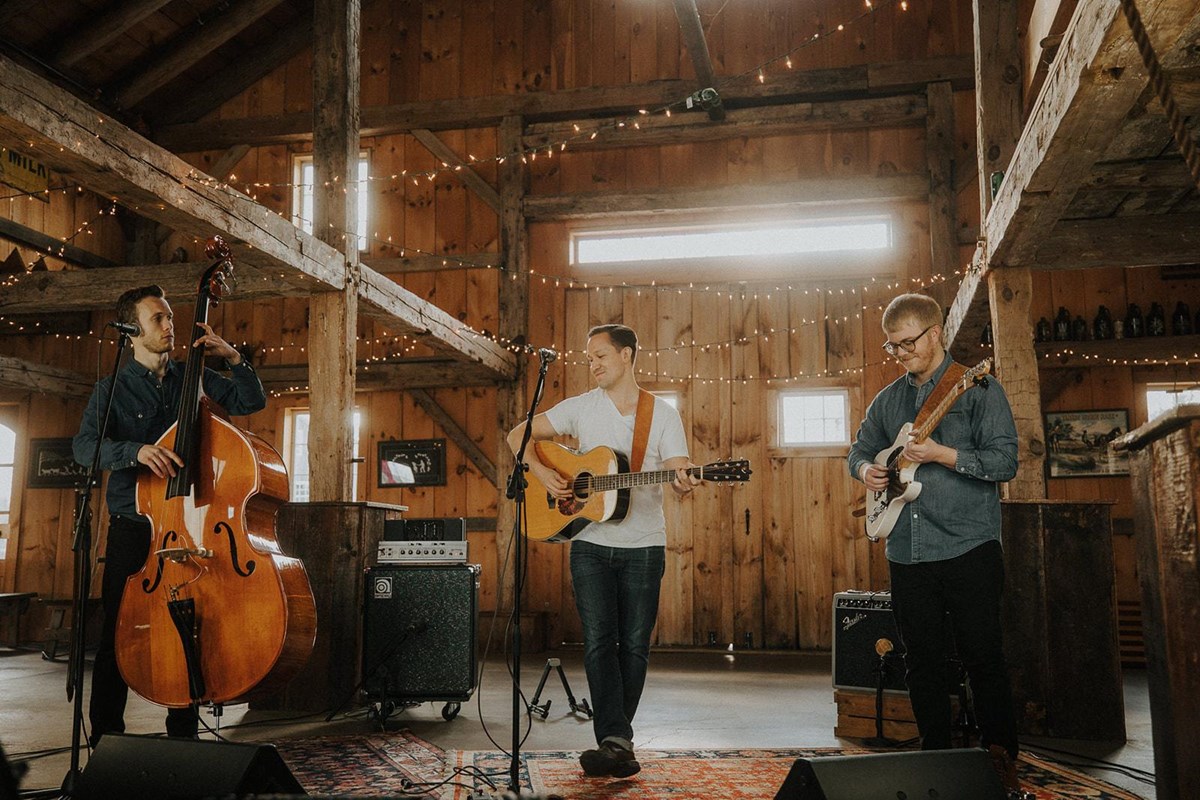 Band of three individuals playing inside a barn. Two play guitars and the third plays the bass.
