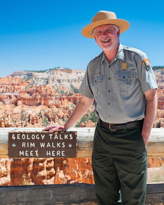 A man in a park ranger uniform stands beside a sign ready Geology Talks meet here