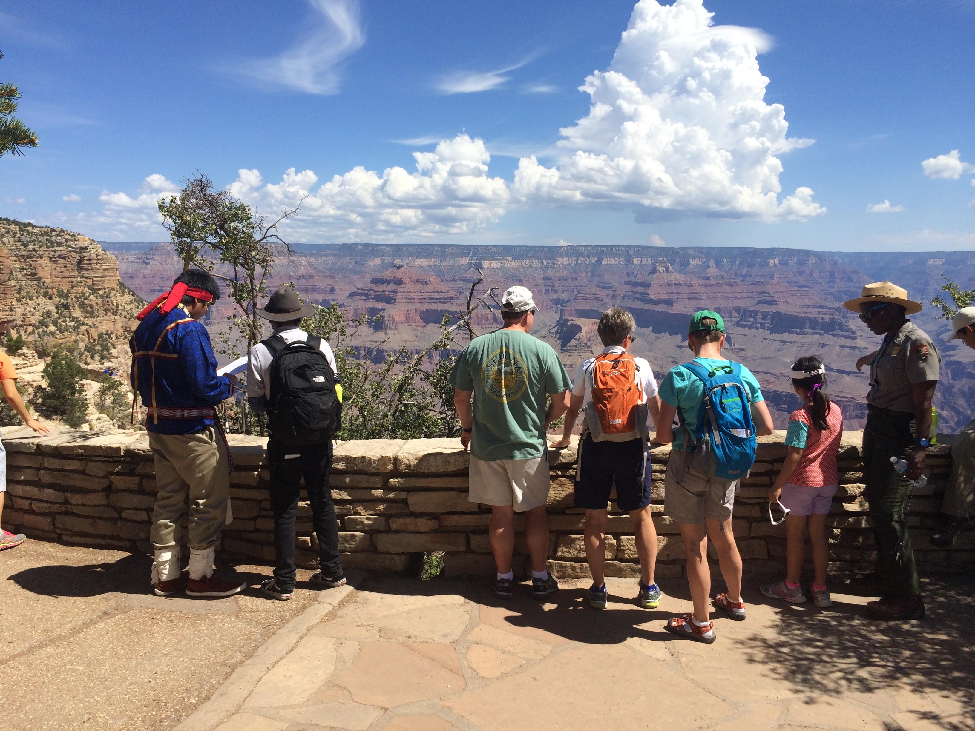 Park rangers and visitors stand along rock wall in front of canyon.