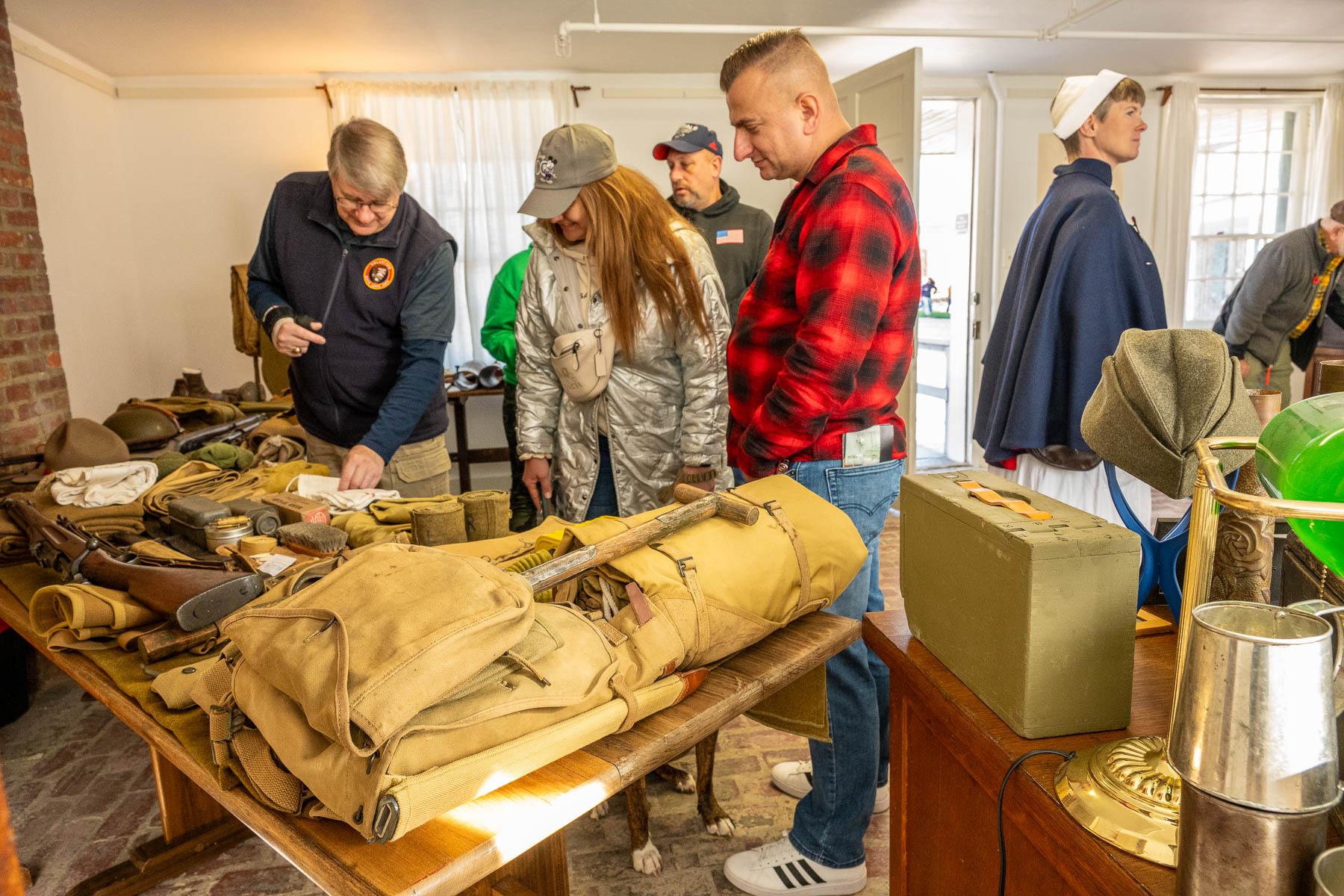 An NPS volunteer in a blue vest shows a man and a woman a table display of WWI era military items.