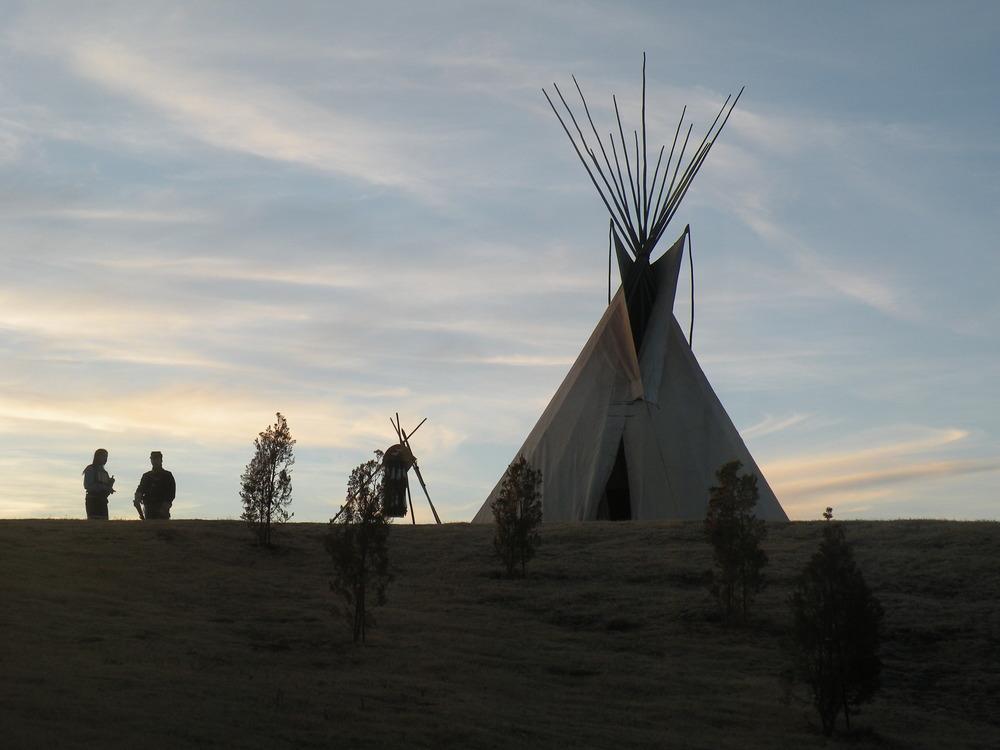 Silhouetted teepee on a hill at dusk with three people standing nearby under a cloudy sky