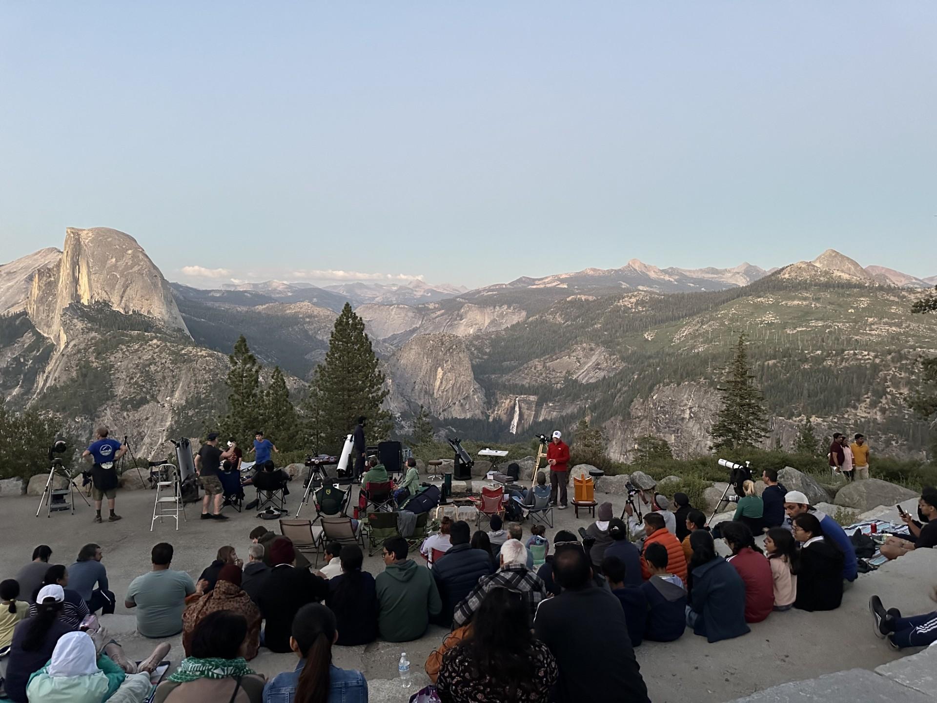 A group of people sits at the Glacier Point amphitheater around sunset, gazing out toward Half Dome.