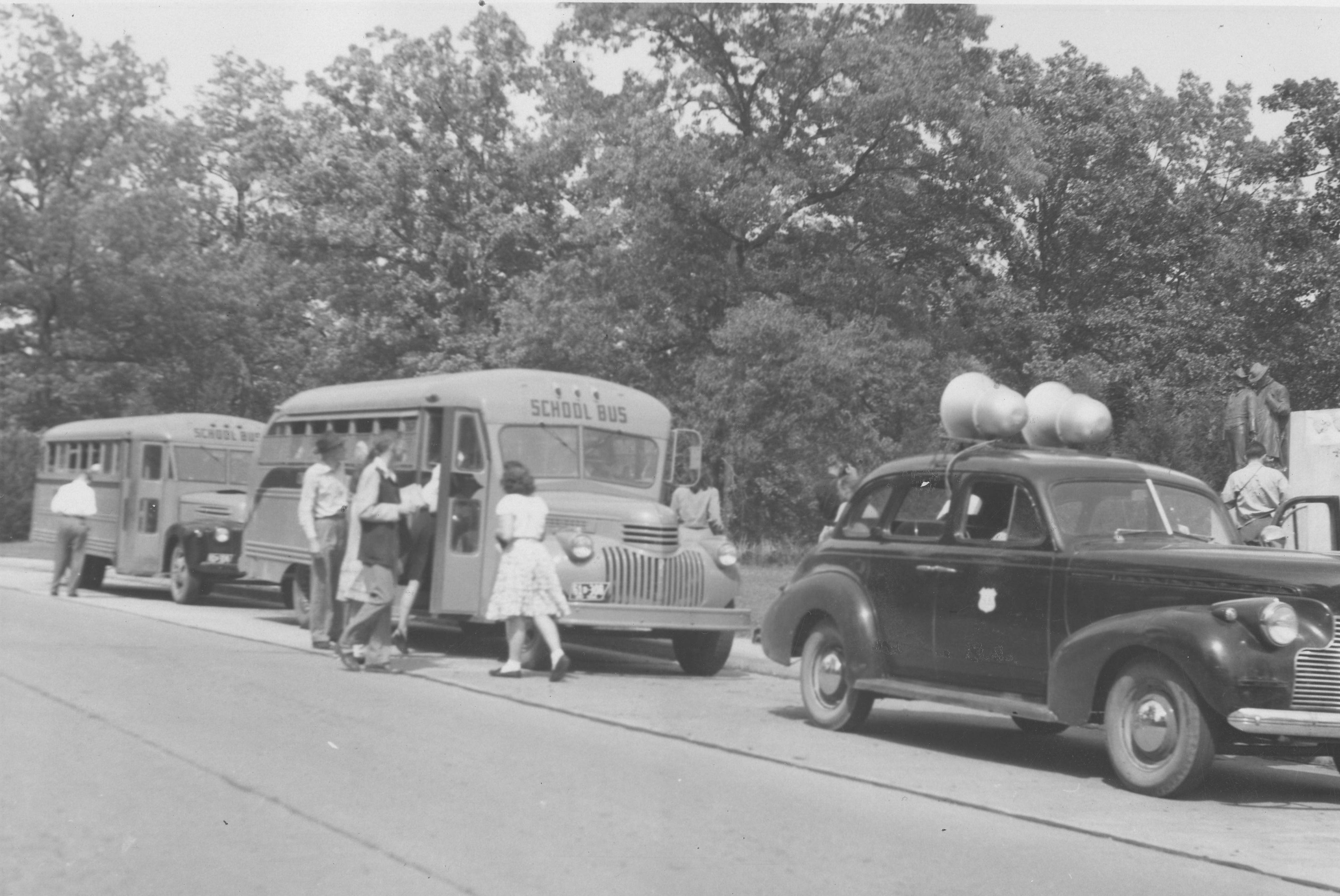 Black-and-white photograph of people boarding two school buses parked along a tree-lined road