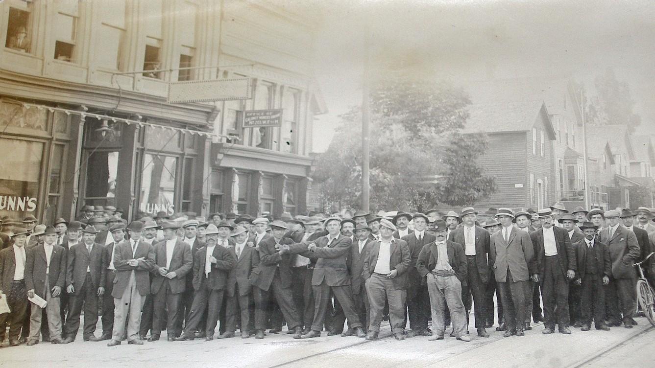 A large group of men pose on the street. Commercial buildings and homes are seen behind them.