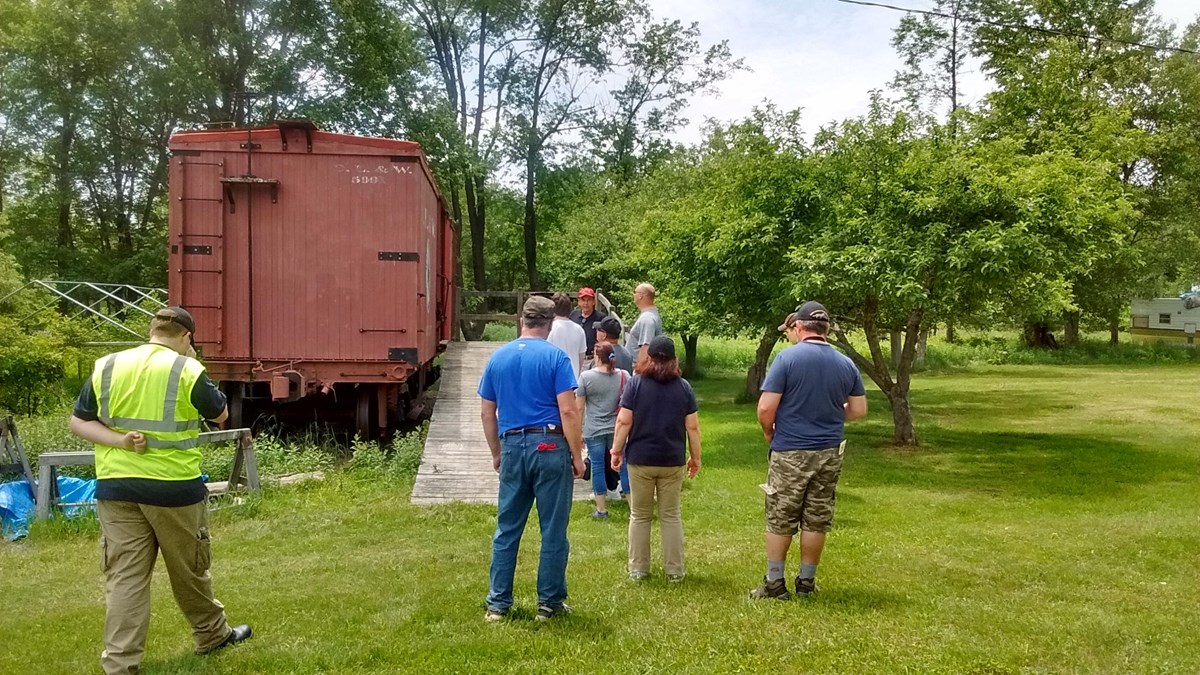 A crowd of approximately 10 people on a guided tour to a box car with a ramp.