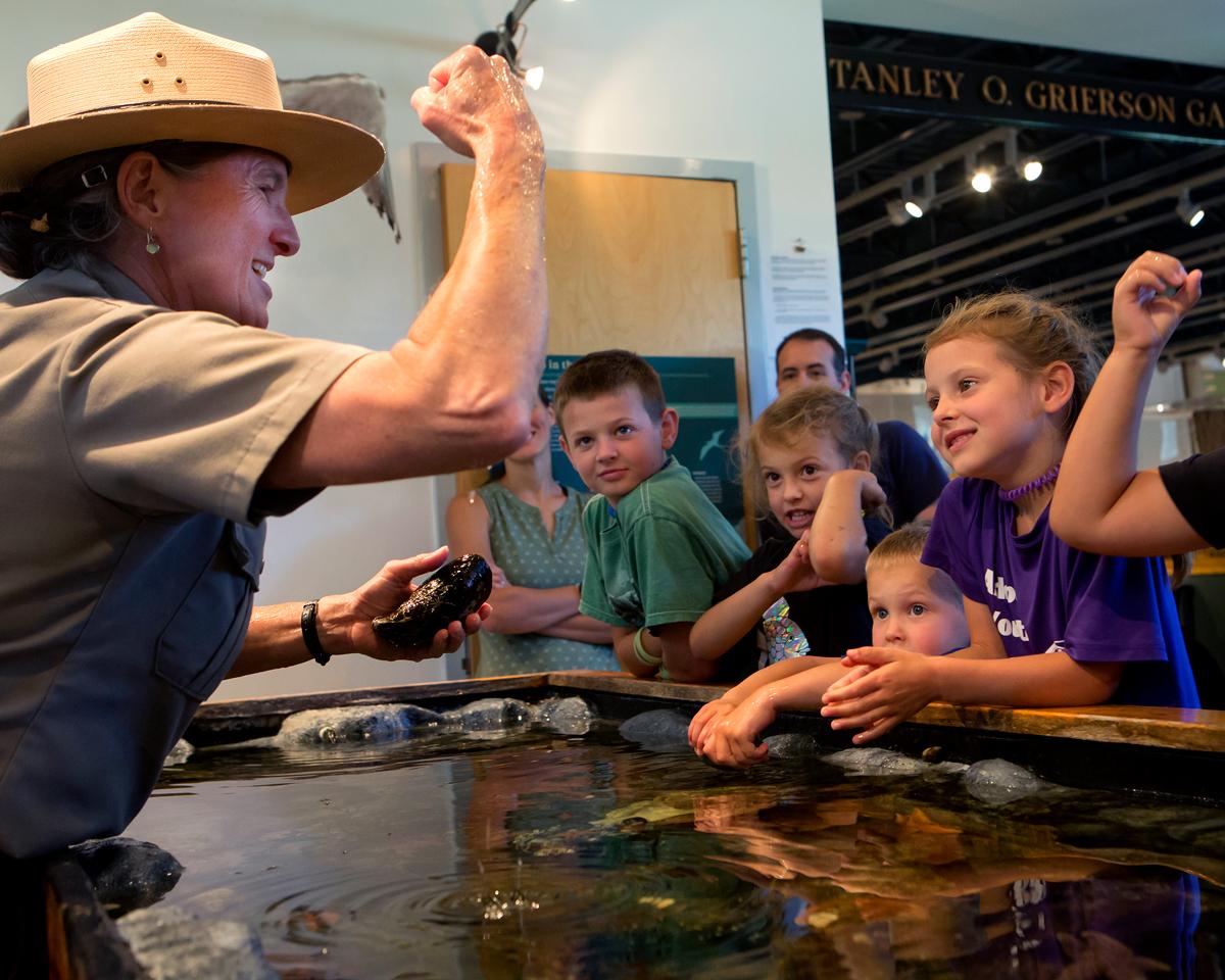 Park ranger in uniform stands by an open water tank with children gathered around the sides