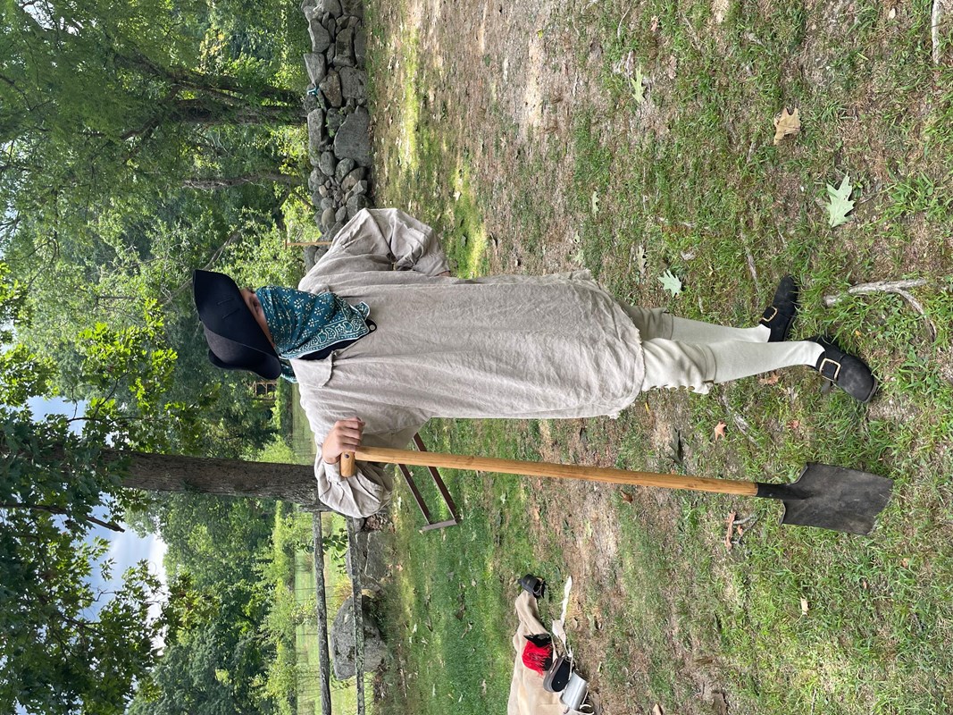 A colonial man with a cloth over his face stands holding a shovel