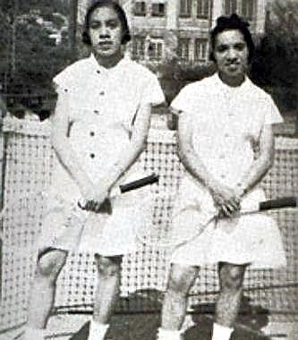 Two African-American women in white tennis attire hold tennis racquets and pose in front of a net.