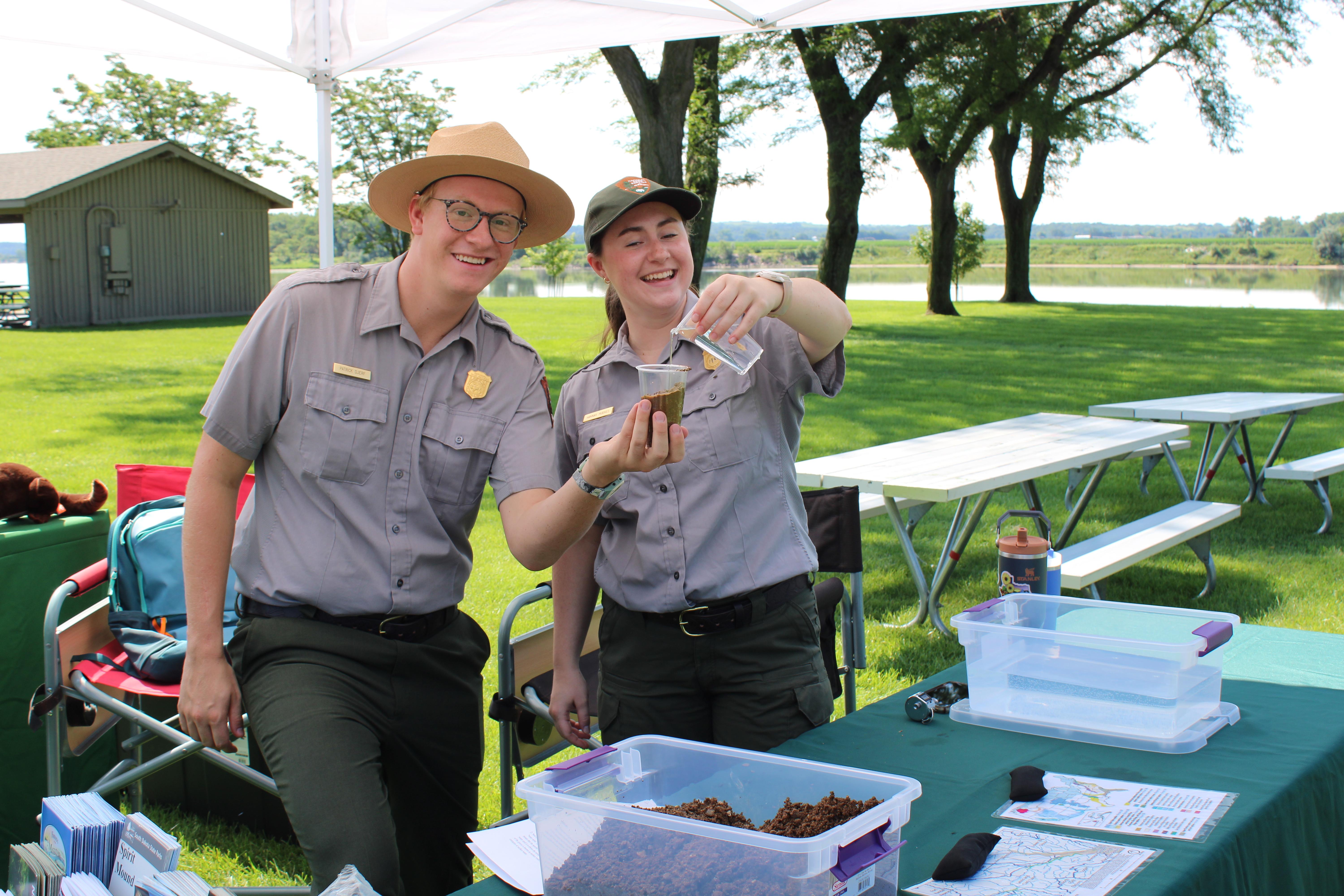 Two rangers a male and a female stand behind two tables doing a demo for junior rangers.