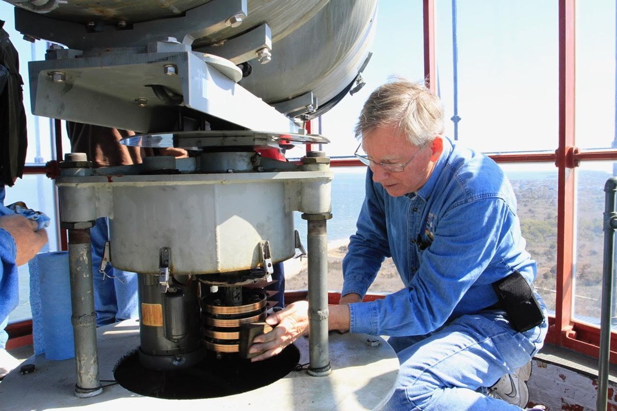 Kneeling man in blue shirt and jeans performs maintenance on lighthouse equipment.