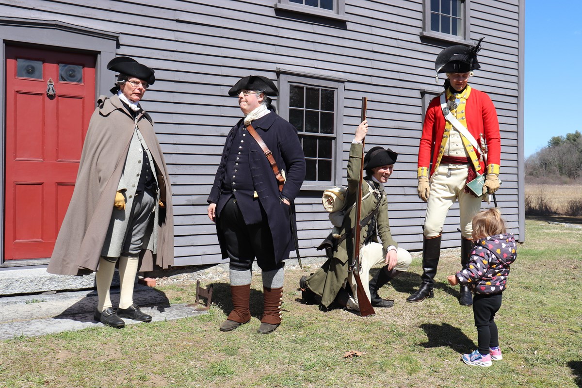 Three Militia and one British reenactor stand in front of a wooden house speaking to a child