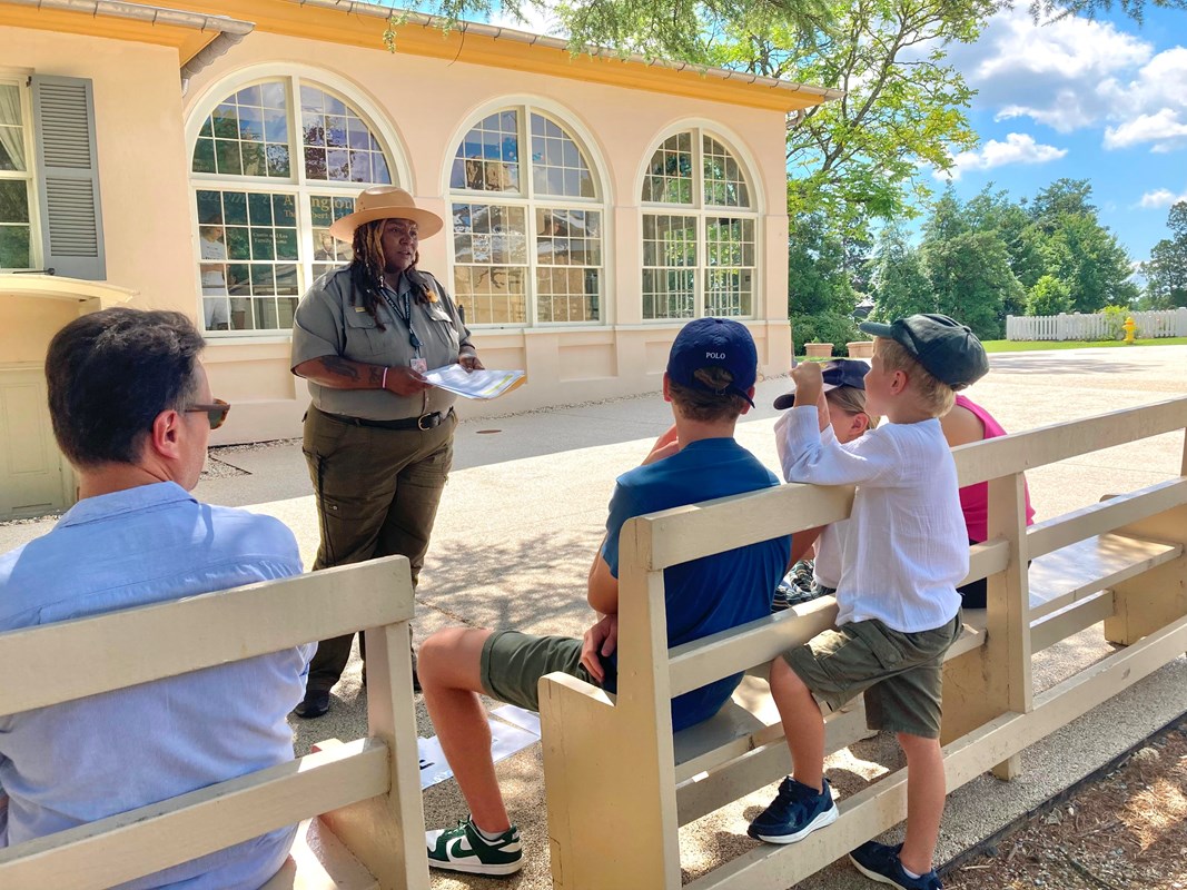 Four visitors sit on a bench. A park ranger stands in front of them showing them a historic image