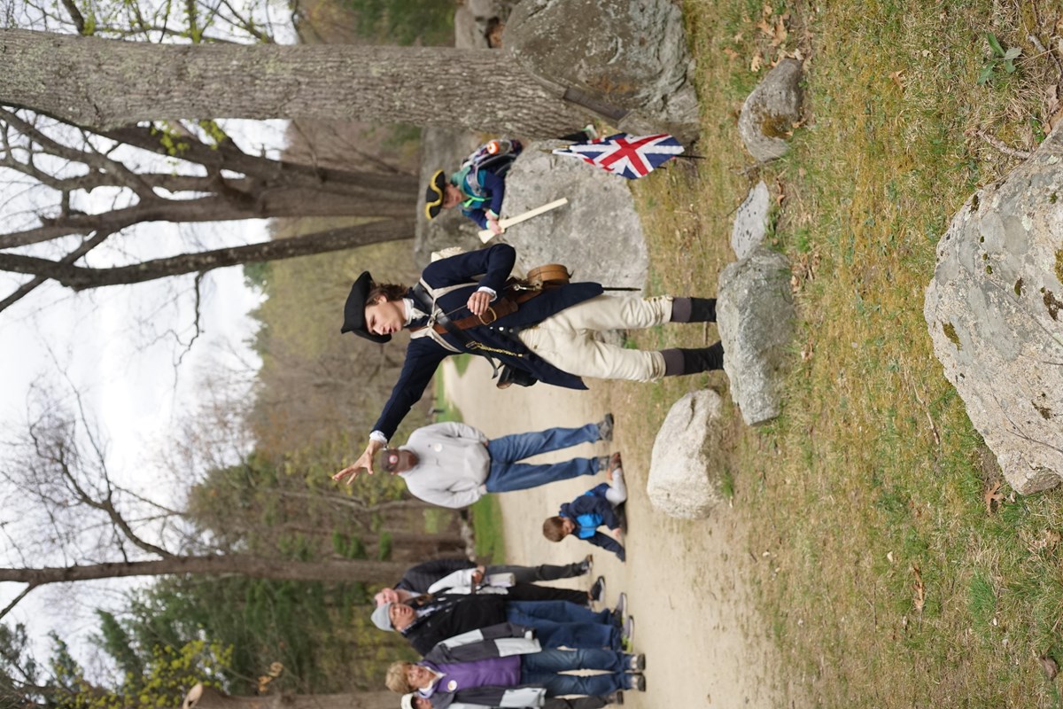 A Park Ranger in 18th Militia clothing stands in front of a crowd talking and pointing.