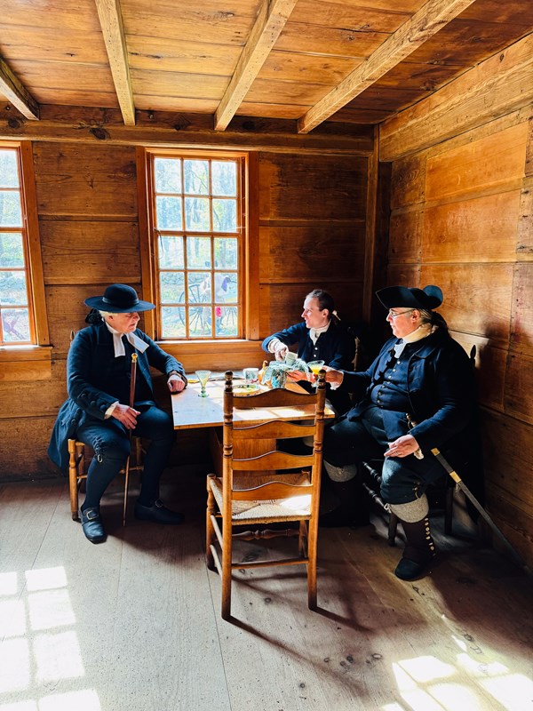 Men in colonial clothing sit near a window in a historic tavern drinking from a punch bowl