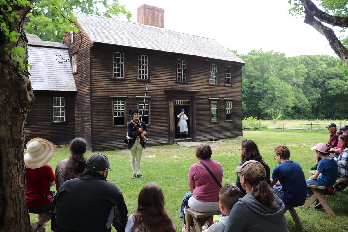 A Park Ranger in militia clothing holds a musket and speaks to a crowd sitting in front of a house.