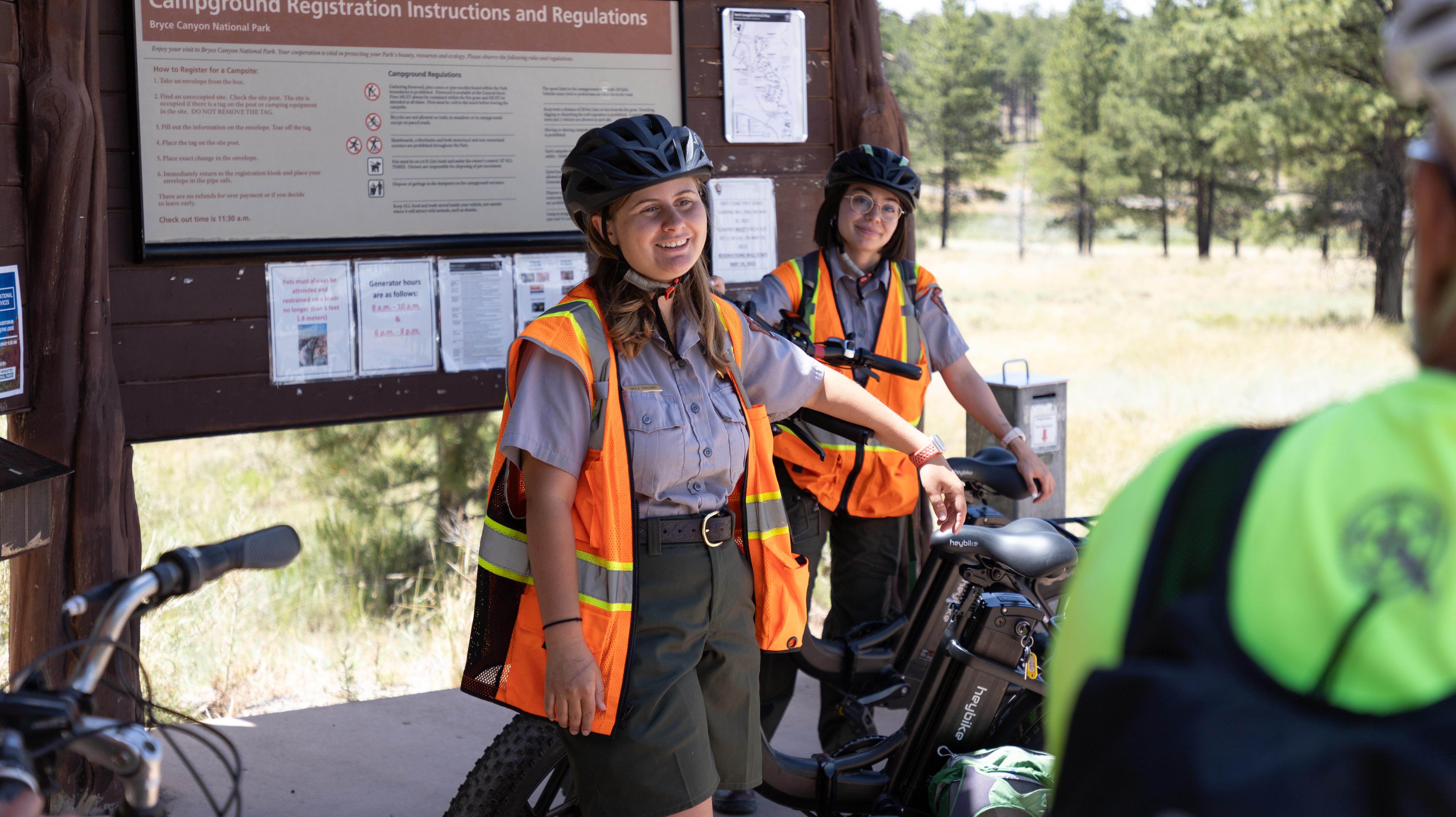 Two park rangers wearing bicycle safety gear speak with a crowd of visitors.