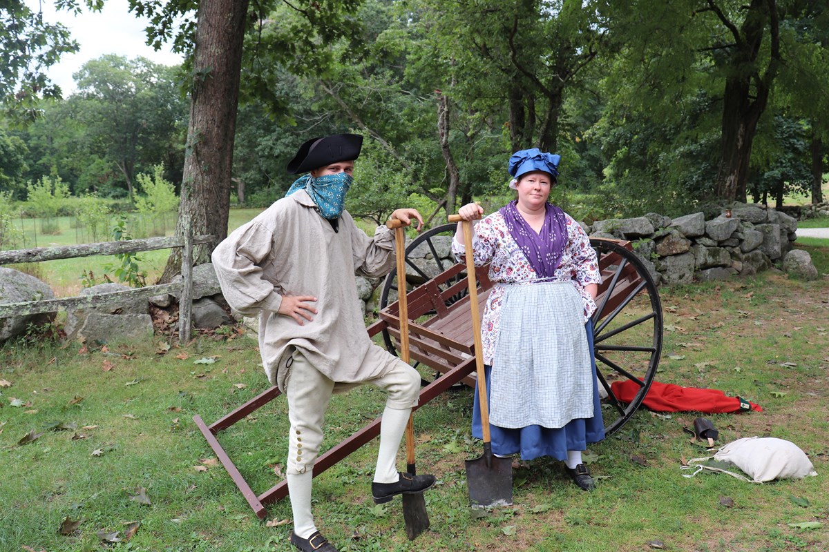 A man and woman in 18th century clothing stand in front of a cart with shovels.