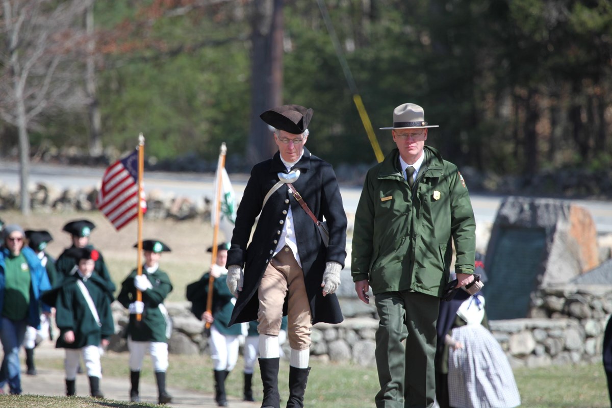 Park Ranger in uniform walks beside a man in a blue colonial coat and black tri-corner hat